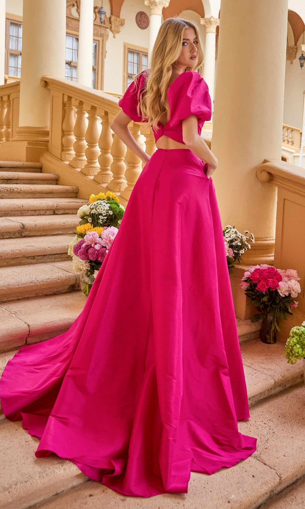 A woman wearing the Puff-Sleeve Long Prom Dress Ava Presley 42269 stands on stone steps lined with flower bouquets, looking over her shoulder.